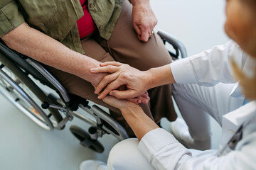 Nurse holding hands with elderly woman in wheelchair during comforting talk.