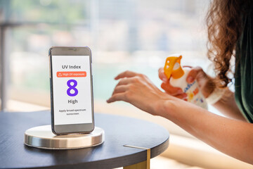 Smartphone with high UV alert in focus, while woman in background applies sunscreen to her skin....