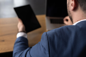 Businessman in suit sitting on orange sofa in office, talking on phone and using tablet and laptop....