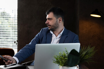 Businessman in suit sitting on orange sofa in office, talking on phone and using tablet and laptop....