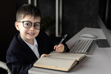 Funny baby boss in glasses sitting at desk with laptop, smartphone, and notebook. Child in suit...