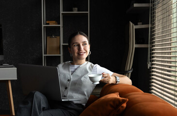 Business woman lying on orange sofa holding coffee cup and laptop. Smiling female in office interior with blinds and sunshine in background.