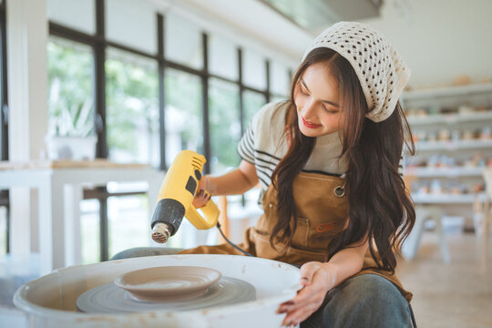 Happy asian woman using handheld dryer to dry clay pottery during ceramic workshop – smiling female enjoying creative hobby, hands-on artistic craft and peaceful leisure activity in studio environment