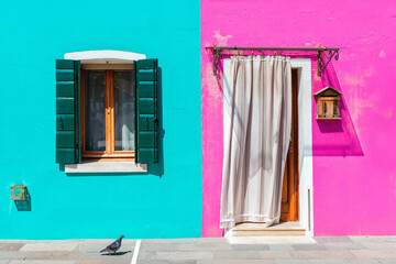 A turquoise house with green-shuttered windows and a fuchsia-colored house with a curtained doorway in Burano, Italy.