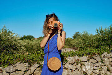 Woman holding orange instant camera and taking photo in natural rural landscape. Girl in blue dress...