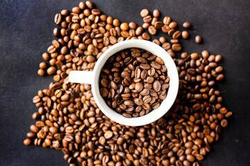 From above close-up view of roasted coffee beans in a white cup on a textured black surface, highlighting the rich textures and colors.