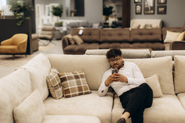 Obraz premium Man Relaxing on Comfortable Couch Using Smartphone in a Modern Furniture Store