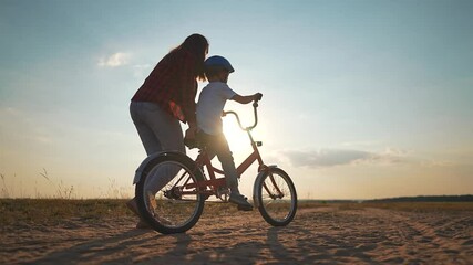 Mother helps son ride bicycle at sunset. Active sport family bonding as mother supports son learn bicycle skills. Outdoor sport Active son practices riding bike with mother guidance in scenic setting - Powered by Adobe