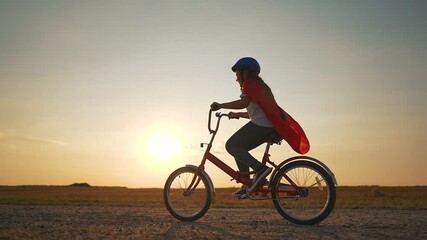 Child superhero cape rides bicycle through park at golden sunset. Joyful child imagines being superhero while pedaling bicycle. Adventure and freedom for happy child on bicycle in scenic park setting.