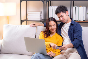 Fototapeta premium Happy asian couple using laptop and credit card for online shopping together in living room at home, man and woman smiling and relax with enjoy a digital lifestyle, convenient payment.
