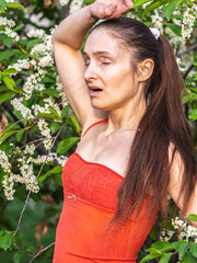 Young woman sneezes in the park against the background of a flowering tree. Allergy to pollen...