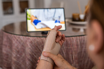 Senior woman wearing wrist brace adjusting it while having online consultation with doctor via tablet, receiving medical advice and support from the comfort of her home