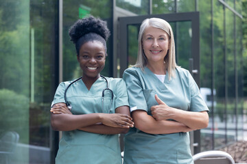 Smiling female doctors in lab coats standing near clinic and looking confident