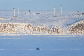 Winter arctic industrial landscape with a wind farm. Wind turbines in the snowy tundra on the coast. Renewable energy in the Arctic. Off-road car is driving on snow and ice. Chukotka, Siberia, Russia.