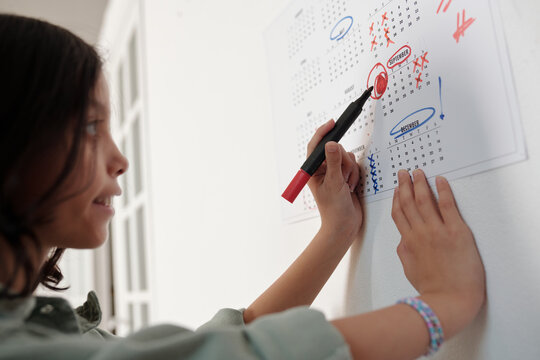 Hispanic girl marking calendar with felt tip pen, preparing for school by highlighting important date, focusing on planning and organization for upcoming academic activities