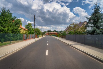 A serene residential street in small village Brzozki in Poland, lined with lush green trees and charming houses, rests under a bright blue sky with fluffy clouds, embodying urban tranquility