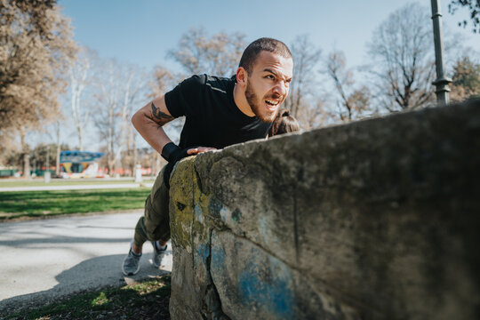 Man engaged in a strenuous parkour move over a concrete barrier, captured in an urban park under bright sunlight. His expression showcases effort and determination.