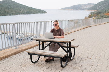 Fototapeta premium Young woman working remotely by the sea with a laptop and solar panel, embracing sustainable travel, mindful lifestyle and digital freedom. Concept of new work, balance and conscious living.