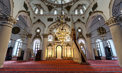 An elaborate chandelier hangs in the center of Hisar Mosque. The historic building has white walls, ornate gold trim, arches, columns, and a red patterned carpet. Located in Izmir, Turkey