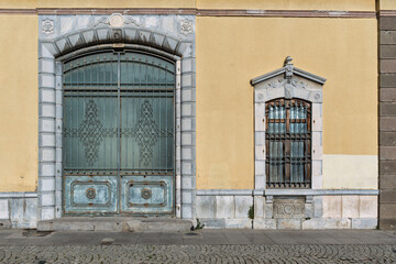 Exterior view of an ornate building facade featuring a large arched gate, and an arched window, yellow wall, and stone details above a cobblestone sidewalk