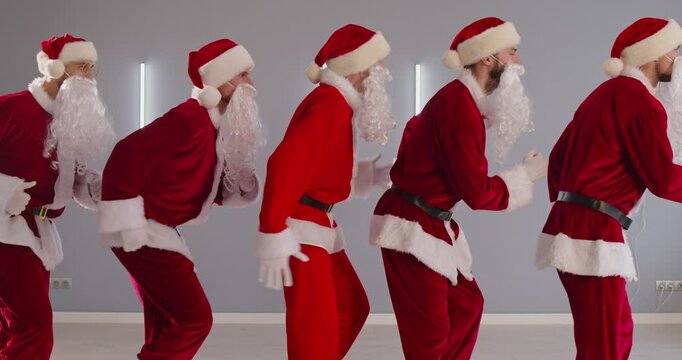 Sideways view of five ridiculous Santa Clauses stepping and moving, standing in row, preparing for Christmas or New Year, wearing red costumes, isolated over grey background. 