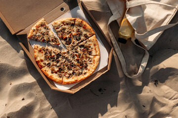 A cozy beach picnic: open pizza box and lemonade in a cotton bag lie on warm sand, glowing in evening sun.