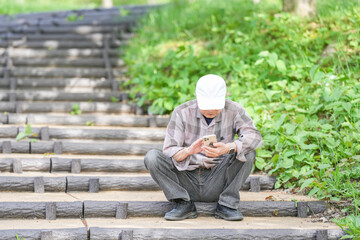 Obraz premium In a lush green park on a sunny May day, a Japanese man in his late 70s, wearing a checkered shirt and white hat, sits on steps using his smartphone. Amid spring foliage, he looks calm and focused.