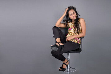 A young woman wearing a floral patterned top, black leggings and black heels, posing with a chair...