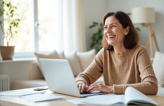 Smiling middle aged woman uses laptop at home. Happy female enjoys hybrid work, online learning or e-business. Lady looks away at copy space smiling. Woman works from home. Indoor. - Powered by Adobe