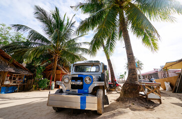 A retro-style jeepney rests beneath lush palm trees on sandy terrain, surrounded by bamboo huts.