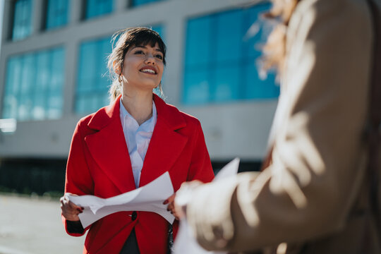 A professional woman in a red coat and white shirt interacts with someone outside a modern office building on a sunny day. She carries documents while confidently engaging in discussion. - Powered by Adobe