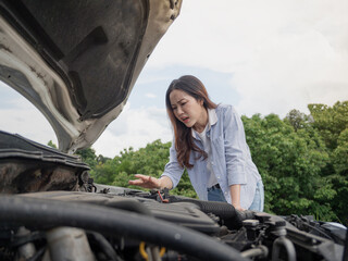A young woman in a jeans shirt looks at the engine under the hood of a broken white car.