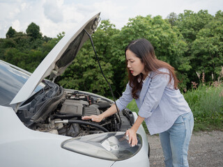 A young woman in a jeans shirt looks at the engine under the hood of a broken white car.