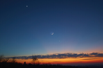 Crescent young Moon with stars, planets and countryside silhouettes.