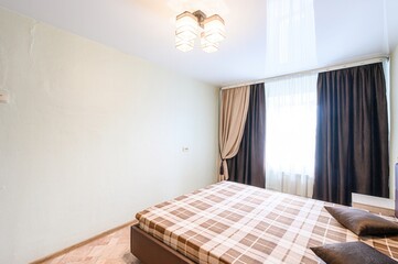 bedroom interior featuring a plaid bed, brown curtains, and a modern ceiling light. The room has cream-colored walls and a shiny stretch ceiling