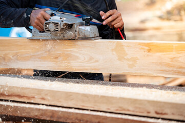 Male carpenter hand using an electric planer on wooden plank at construction site. Woodworker planing timber in his workshop with sawdust spreading around. Lumber industry and carpentry work tool