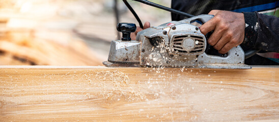 Male carpenter hand using an electric planer on wooden plank at construction site. Woodworker planing timber in his workshop with sawdust spreading around. Lumber industry and carpentry work tool