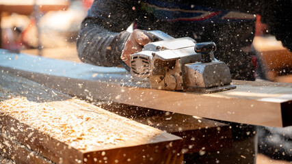 Male carpenter hand using an electric planer on wooden plank at construction site. Woodworker planing timber in his workshop with sawdust spreading around. Lumber industry and carpentry work tool