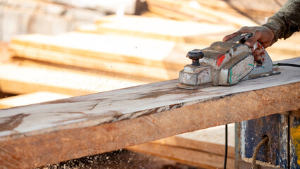 Male carpenter hand using an electric planer on wooden plank in workshop. Woodworker planing timber at construction site with sawdust spreading around. Work tool for carpentry. Lumber industry concept