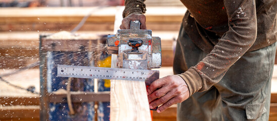 Male carpenter hand using angle ruler for wood squaring and planing on wooden plank by electric planer on workbench in workshop. Wood measuring and shaving process for lumber industry