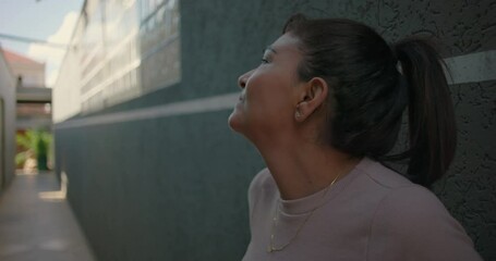 Latina woman leans against wall with hand on forehead and places hands on hips in visible distress, expressing physical discomfort and emotional fatigue in alley