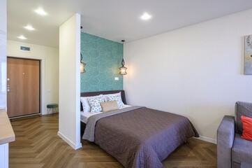 cozy bedroom with a bed featuring patterned pillows and a brown quilt, flanked by hanging lamps. The room includes a green patterned accent wall and light wood floors