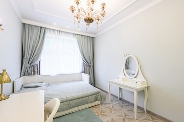 well-lit bedroom features a bed by a window with light blue curtains, a vanity with a mirror, and a decorative chandelier. The room is clean and has a classic, elegant style