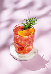 Red orange cocktail with ice and rosemary on a marble stand on a pink background