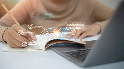 A young woman is studying online with a laptop and notebook. She is taking notes while using...