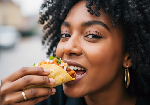 A close-up portrait of a smiling young Black woman with curly hair, happily taking a bite out of a fresh taco filled with meat and salsa
