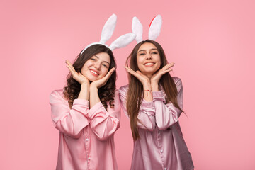 Two cheerful young women wearing matching satin pink pajamas and bunny ears headbands on a pastel...