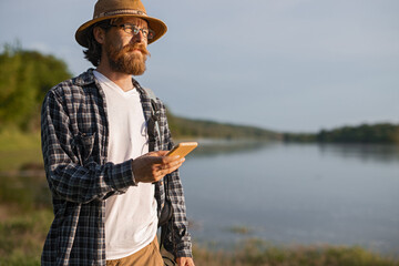 A man wearing casual attire and glasses, carrying a phone, admires the serene view by the lakeside as the sun sets, portraying themes of exploration, mindfulness, and connection with nature.