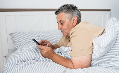 Happy mature grey hair woman with smartphone laying in a bed in the morning.