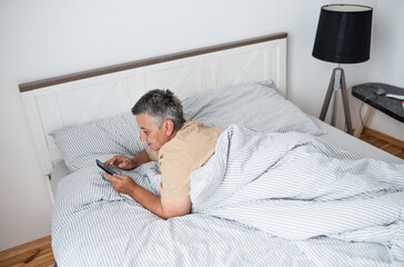 Happy mature grey hair woman with smartphone laying in a bed in the morning.